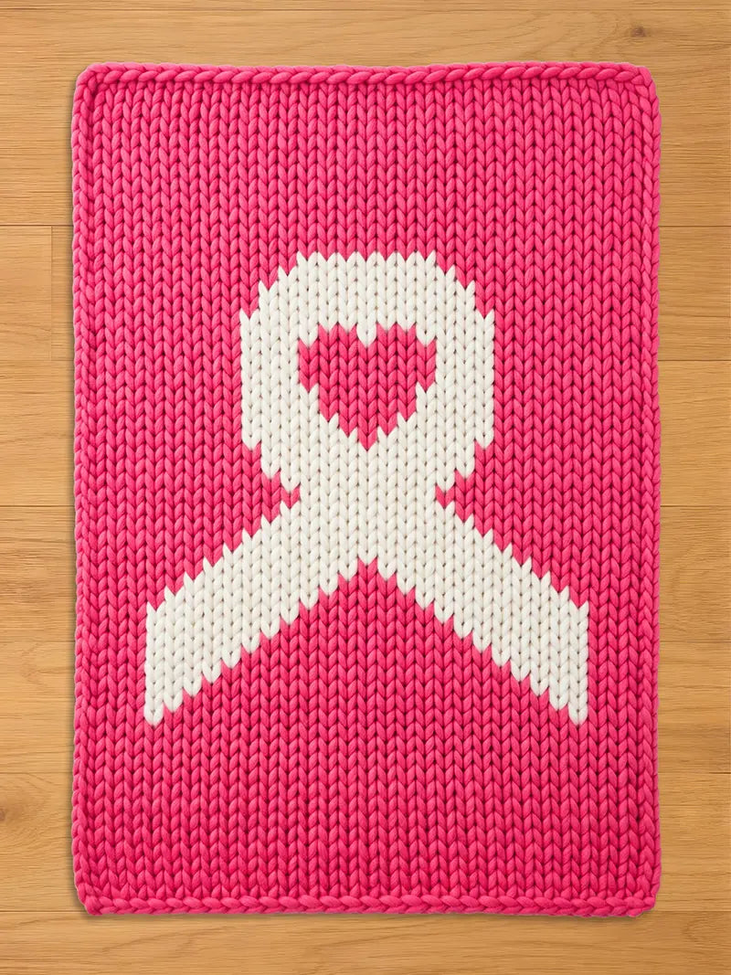 Bright pink chunky knit blanket featuring a large white awareness ribbon design in the center, with a matching pink heart inside the top loop of the ribbon