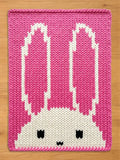 A pink and white knitted bunny blanket. The blanket features a large bunny face with long ears, black eyes, and a small mouth. The background is a solid pink color, and the bunny is white.