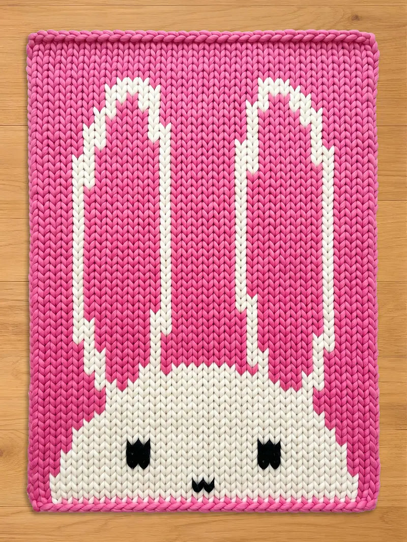 A pink and white knitted bunny blanket. The blanket features a large bunny face with long ears, black eyes, and a small mouth. The background is a solid pink color, and the bunny is white.