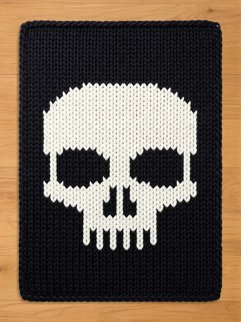 A black chunky knit blanket featuring a large, contrasting white skull design in the center, laid flat against a light wooden surface