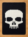 A black chunky knit blanket featuring a large, contrasting white skull design in the center, laid flat against a light wooden surface