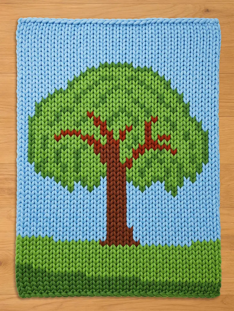 A knitted blanket featuring a tree design. The tree has a brown trunk and branches with a green canopy. The background is blue, with green at the base, creating a landscape scene. The blanket is made of soft yarn.