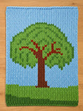 A knitted blanket featuring a tree design. The tree has a brown trunk and branches with a green canopy. The background is blue, with green at the base, creating a landscape scene. The blanket is made of soft yarn.