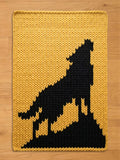 A rectangular, knitted chunky blanket featuring a black silhouette of a howling wolf against a yellow background. The blanket has a thick, textured appearance, with the wolf design prominently displayed. The blanket is suitable for home decor.