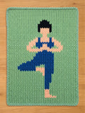 A rectangular chunky knit blanket laid flat. The blanket features a chunky mint green background with a geometric, pixel-art style pattern of a person with black hair, wearing a blue sports bra and leggings, balancing on one leg with hands clasped in the yoga tree pose.