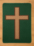 A knitted green blanket featuring a large cross design. The cross is tan with a brown border, centered on the blanket. The blanket's texture is visible, with a thick, woven appearance. The overall design is simple and geometric.