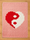 A pink knitted blanket featuring a red and white yin-yang heart design. The blanket has a thick, textured appearance and a soft, pastel color palette. The design is centered on the blanket.