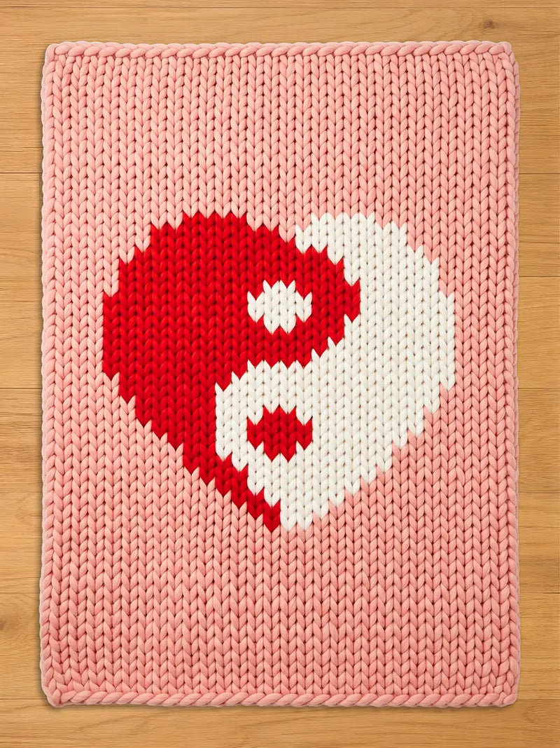 A pink knitted blanket featuring a red and white yin-yang heart design. The blanket has a thick, textured appearance and a soft, pastel color palette. The design is centered on the blanket.