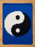 A chunky knit blanket featuring a large black and white yin-yang symbol centered on a vibrant blue background, laid flat on a wooden surface.