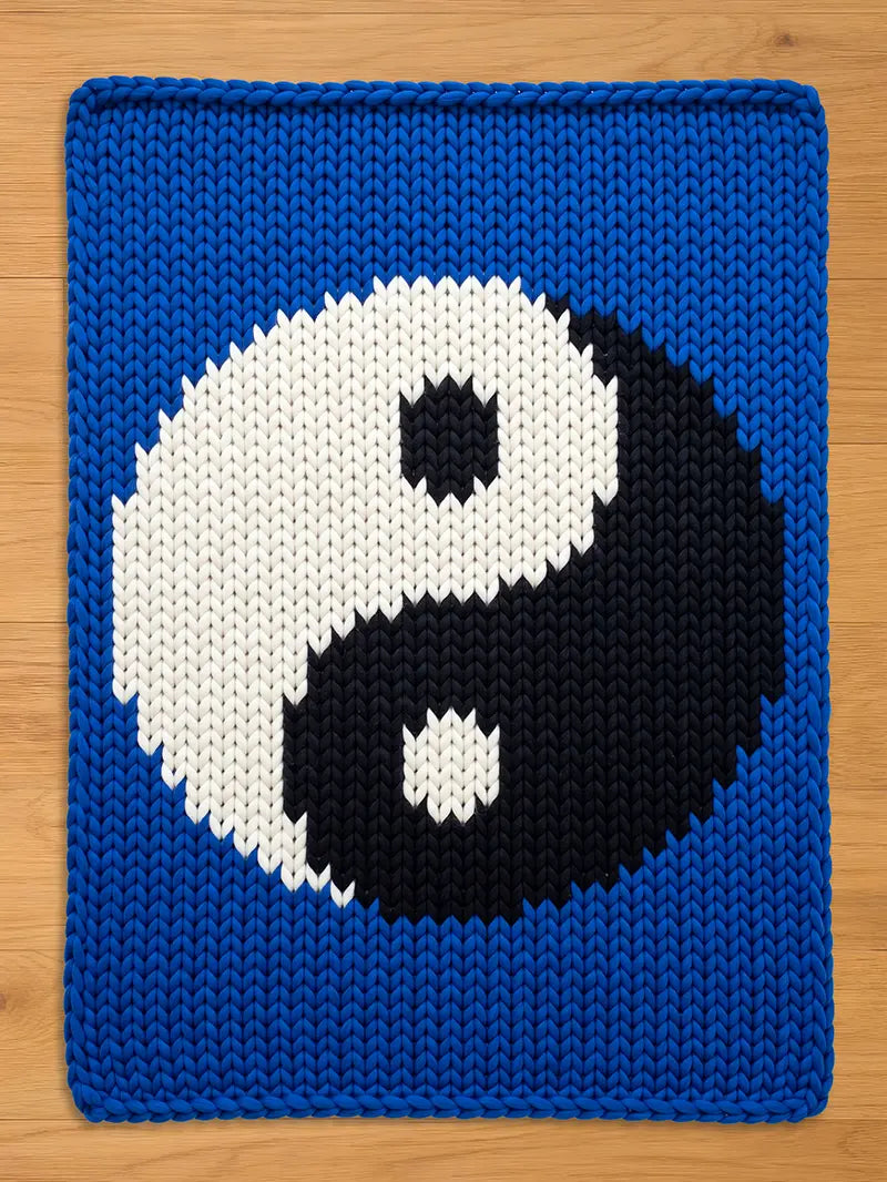 A chunky knit blanket featuring a large black and white yin-yang symbol centered on a vibrant blue background, laid flat on a wooden surface.