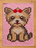 A chunky knit blanket featuring a knitted pattern of a brown and tan Yorkshire Terrier dog with a red bow on its head, sitting against a solid light pink background