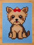 A rectangular chunky knit blanket laid flat on a wooden surface. The blanket has a light blue background and features a large, pixel-style knitted design of a sitting Yorkshire Terrier. The dog has tan, dark brown, and black fur, with a bright red bow on top of its head
