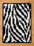 A rectangular, hand-knitted chunky blanket with a bold, geometric pattern. The design features alternating bands of black and white, creating a striking visual contrast. The blanket's edges are neatly finished, and the overall aesthetic is modern and graphic.