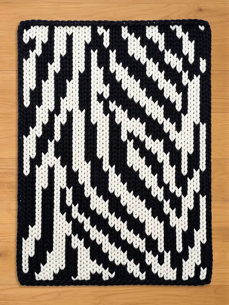 A rectangular, hand-knitted chunky blanket with a bold, geometric pattern. The design features alternating bands of black and white, creating a striking visual contrast. The blanket's edges are neatly finished, and the overall aesthetic is modern and graphic.