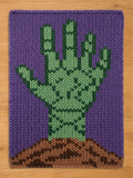 A rectangular knitted blanket featuring a graphic design of a green hand emerging from brown soil against a solid purple background. The knit texture is visible throughout the piece, showcasing a clear contrast between the colors.