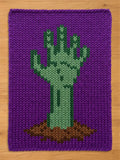 A rectangular purple knitted blanket featuring a central graphic of a green hand emerging from a patch of brown soil. The hand is depicted in varying shades of green, with a distinct thumb and four fingers reaching upward.