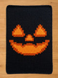 A black chunky knit blanket featuring a vibrant orange knitted jack-o'-lantern face with triangular eyes, a nose, and a wide, jagged smile